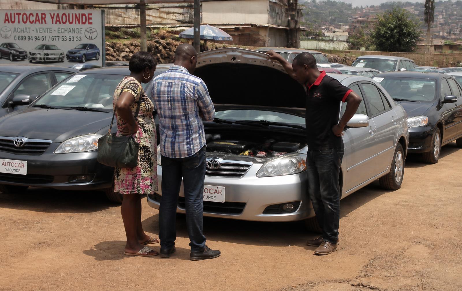 Parc automobile à Yaoundé : acheteurs et vendeur inspectant un véhicule avec le capot ouvert